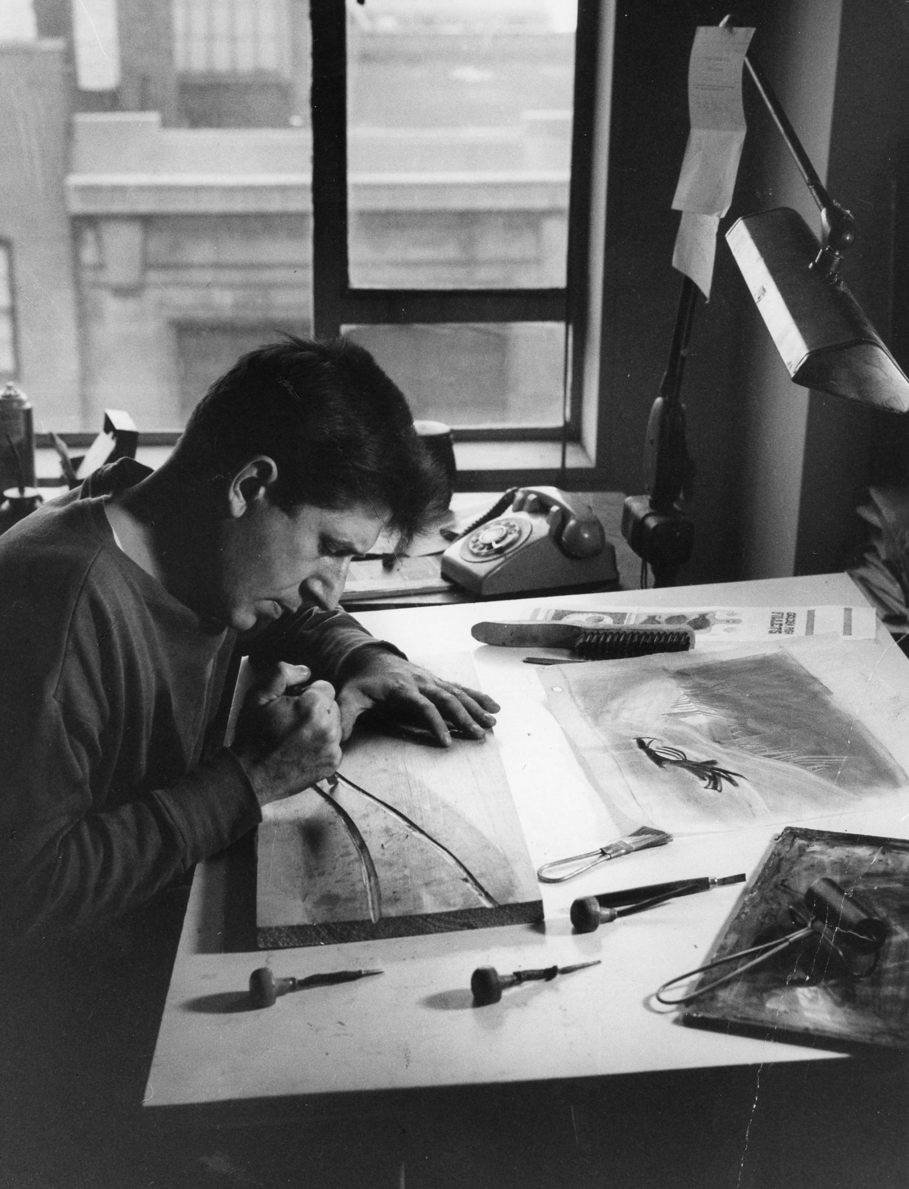 Black and white photo of Walter Ferro in his studio carving a second of two curving lines into a block of wood atop a drafting table with rotary phone and window with brick buildings in the background